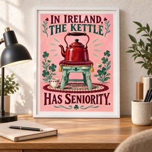 May include: A framed art print featuring a red kettle resting on a small, light blue stool with a cushion. The background is pink with green shamrock illustrations. The text reads: IN IRELAND, THE KETTLE HAS SENIORITY. A small white cup sits nearby.
