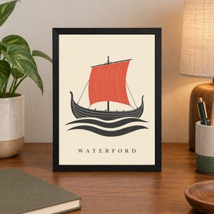 Pode incluir: Uma impress&atilde;o art&iacute;stica emoldurada apresentando um navio viking preto estilizado com uma grande vela vermelha, navegando sobre ondas pretas em um fundo creme. A palavra WATERFORD est&aacute; impressa em letras mai&uacute;sculas pretas na parte inferior.