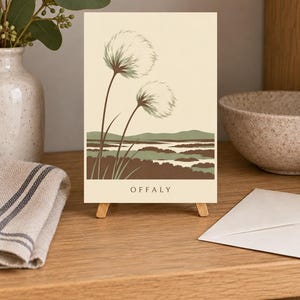 May include: A rectangular greeting card featuring a cream background with a landscape illustration of two tall, fluffy seed heads in the foreground and rolling hills in the distance. The color palette includes shades of sage green and brown. The text OFFALY is printed in black at the bottom.