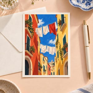 Op de afbeelding: Een rechthoekige kunstdruk met een levendig mediterraan straatbeeld met hoge oranje en gele gebouwen. Wasgoed hangt aan lijnen tussen de structuren tegen een helderblauwe lucht. Potplanten sieren balkons en een verre koepel is zichtbaar.