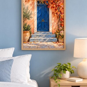 May include: A framed wall art print depicting a vibrant blue door set into a peach-coloured wall. The scene features stone steps, a potted olive tree, and autumn foliage in shades of red and orange cascading over the doorway entrance.