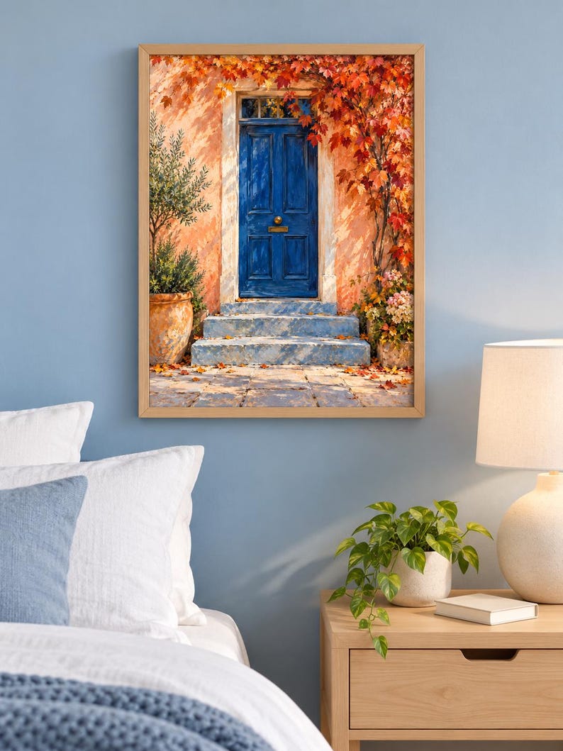 May include: A framed wall art print depicting a vibrant blue door set into a peach-coloured wall. The scene features stone steps, a potted olive tree, and autumn foliage in shades of red and orange cascading over the doorway entrance.