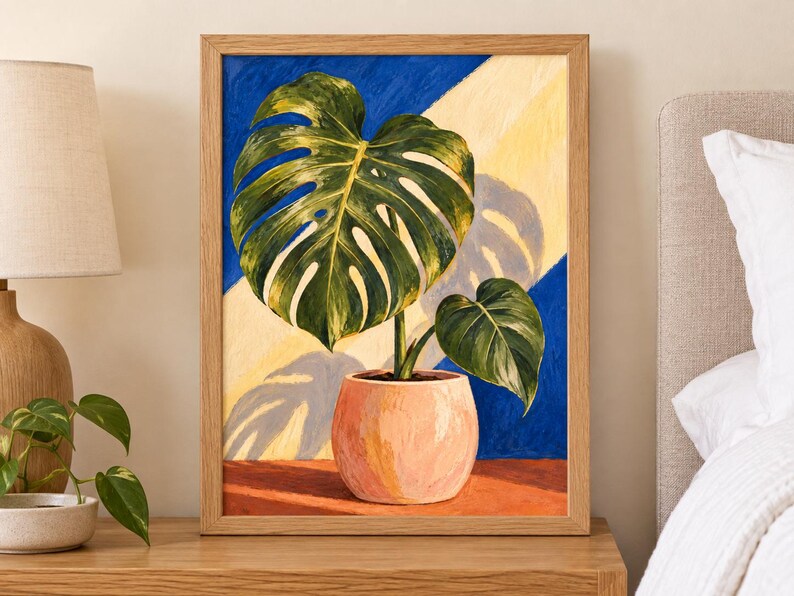 Puede incluir: Un cuadro enmarcado de una planta monstera verde en una maceta color melocot&oacute;n. El fondo presenta rayas diagonales azules y amarillas, con una sombra oscura proyectada sobre la superficie. La obra est&aacute; en un marco de madera clara.