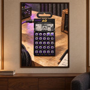 May include: A black Teenage Engineering PO-20 Arcade pocket operator synthesizer featuring a digital LCD screen, a grid of small square buttons with purple accents, two rotary knobs, and a purple audio cable plugged into the side port.
