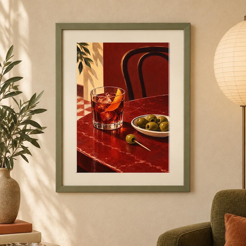 May include: A framed art print depicting a glass of dark amber liquid with ice and an orange peel garnish on a deep red table. Next to the glass is a small white bowl containing green olives and a single olive on a toothpick on the table surface.