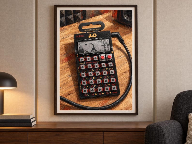 May include: A black Teenage Engineering PO-28 robot pocket operator synthesiser featuring a monochrome LCD screen, a grid of silver buttons with red accents, and a black audio cable plugged into the top right corner, all framed in a dark wood frame.