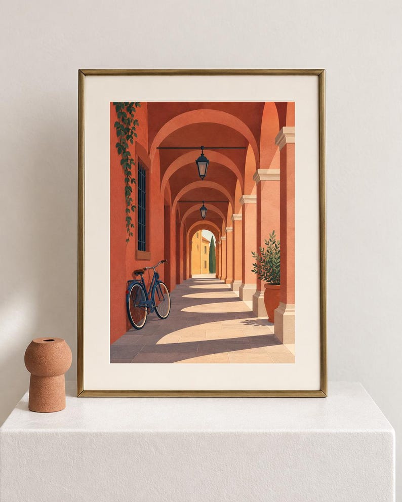 Pu&ograve; includere: Una stampa artistica incorniciata che raffigura un porticato color terracotta con una bicicletta blu appoggiata a un pilastro. La scena presenta lanterne sospese, una pianta in vaso e piante rampicanti, il tutto in una cornice dorata.