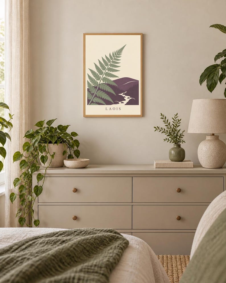 May include: A rectangular wall art print in a light wood frame featuring a green fern frond overlapping a purple mountain landscape with a winding river and the text LAOIS printed in black at the bottom centre of the cream-coloured background.