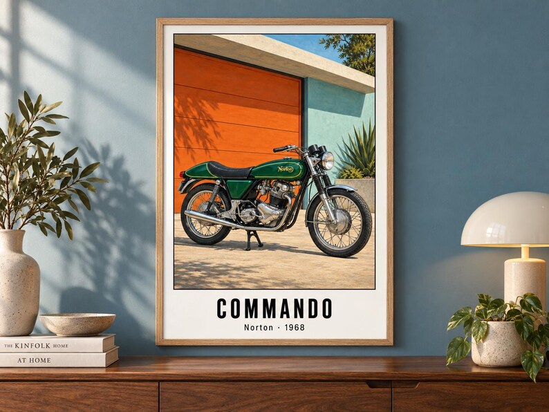 May include: A framed wall art print featuring a green Norton Commando motorcycle parked in front of an orange garage door. The text at the bottom reads COMMANDO Norton 1968 in black capital letters against a white background within the print.