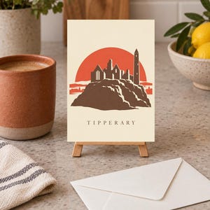 May include: A rectangular greeting card featuring a graphic illustration of the Rock of Cashel in Tipperary, Ireland. The design displays a brown stone castle silhouette against a large orange sun on a cream background, with the word TIPPERARY below.