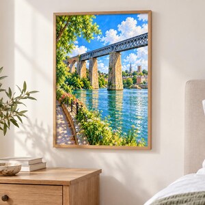 May include: A framed wall art print depicting a stone and metal railway bridge spanning a blue river. The scene includes lush green trees, a stone embankment with colorful flowers, and a bright blue sky with white clouds above a distant town.