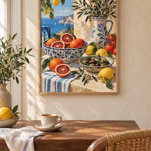 Op de afbeelding: Een ingelijste kunstdruk met een mediterrane kustsc&egrave;ne, voorzien van een blauw-wit patroonkom gevuld met bloedsinaasappels, een bijpassende kan met olijftakken, een kom met olijven en citroenen op een blauw-wit gestreepte doek.