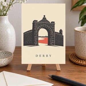 May include: A rectangular print on a small wooden easel featuring a black graphic illustration of a stone archway and city walls. The word DERRY is printed in black capital letters below the arch, with a red path visible through the opening.