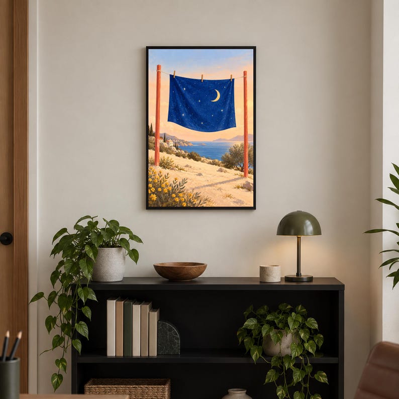 May include: A framed wall art print depicting a dark blue fabric with a crescent moon and stars hanging on a clothesline between two red poles. The scene features a coastal landscape with a sandy foreground, yellow flowers, and a blue sea.