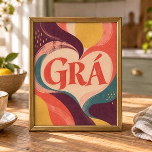 May include: A rectangular framed print featuring the word GR&Aacute; in bold red letters centred within a cream-coloured heart shape. The background consists of abstract, flowing shapes in shades of yellow, red, dark purple, and teal with small yellow dashes.