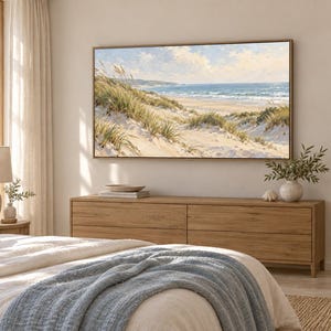 May include: A rectangular landscape painting featuring a coastal scene with sand dunes, tall beach grass, and a blue ocean under a cloudy sky, displayed in a thin wooden frame on a light-coloured wall above a long wooden dresser in a bedroom.