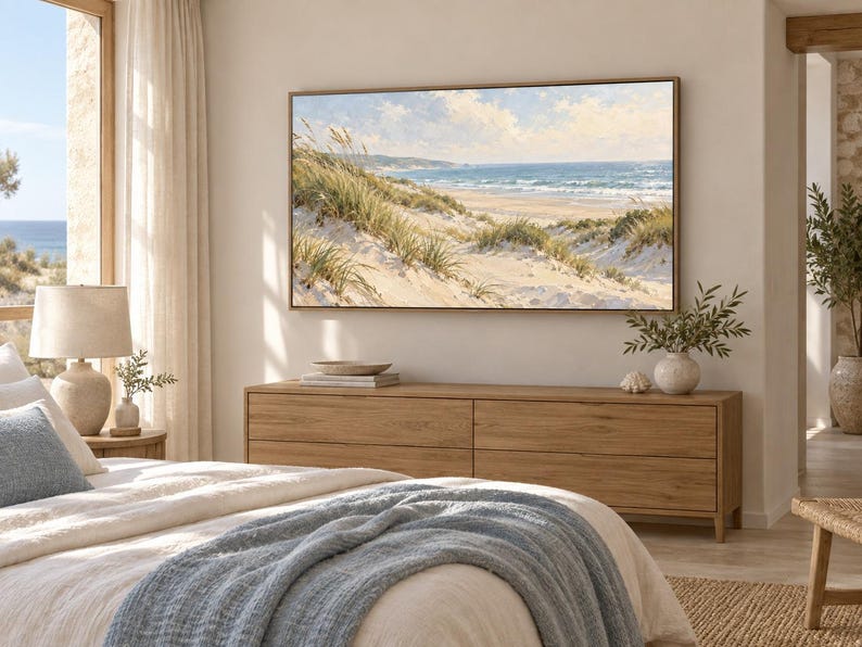May include: A rectangular landscape painting featuring a coastal scene with sand dunes, tall beach grass, and a blue ocean under a cloudy sky, displayed in a thin wooden frame on a light-coloured wall above a long wooden dresser in a bedroom.