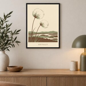 May include: A framed wall art print featuring a cream background with two stylized dandelion-like plants in the foreground. Below the plants is a landscape of green and brown rolling hills and water. The text OFFALY is printed at the bottom.
