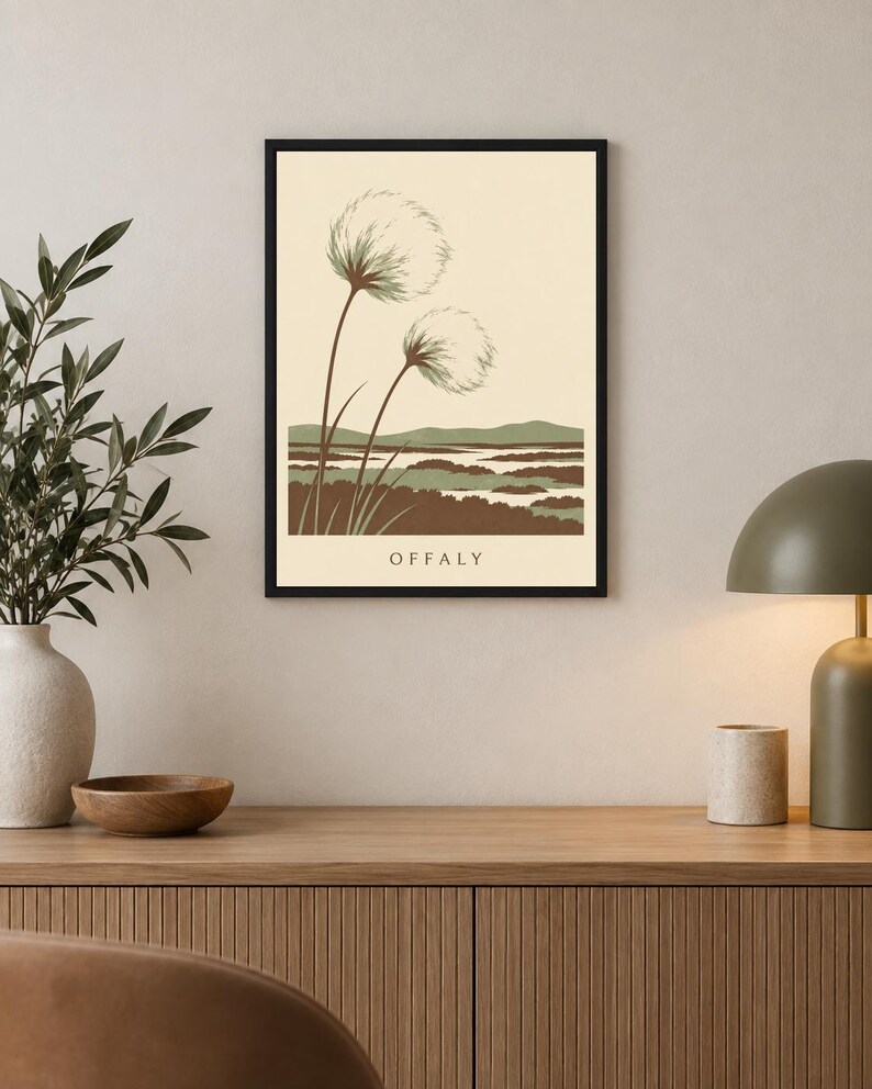 May include: A framed wall art print featuring a cream background with two stylized dandelion-like plants in the foreground. Below the plants is a landscape of green and brown rolling hills and water. The text OFFALY is printed at the bottom.