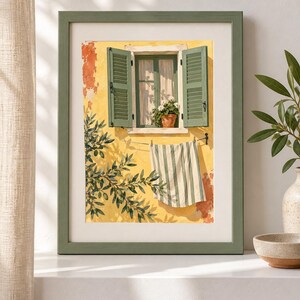 May include: A framed wall art print depicting a yellow stucco wall with an open window featuring green shutters and a potted plant. A striped towel hangs on a clothesline below the window, and olive tree branches are visible in the foreground.