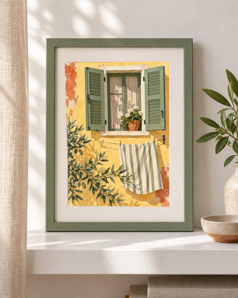 May include: A framed wall art print depicting a yellow stucco wall with an open window featuring green shutters and a potted plant. A striped towel hangs on a clothesline below the window, and olive tree branches are visible in the foreground.