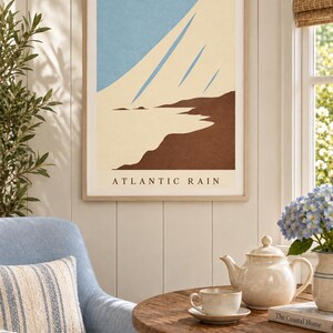 May include: A framed wall art print titled ATLANTIC RAIN featuring a minimalist landscape illustration with a light blue sky, cream-colored diagonal streaks, and a dark brown coastline. The artwork is displayed in a light wood frame.