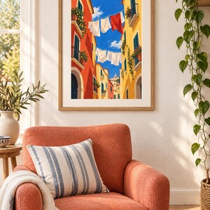 Op de afbeelding: Een ingelijste kunstdruk toont een smalle straat tussen hoge oranje en gele gebouwen met groene luiken en balkons. Wasgoed hangt aan lijnen die onder een helderblauwe lucht zijn gespannen, met witte en rode stoffen.