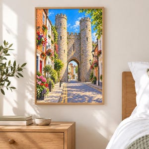 Op de afbeelding: Een ingelijste wandkunstprint met een middeleeuws stenen poortgebouw met twee torens, geflankeerd door bakstenen gebouwen versierd met kleurrijke hangende bloemenmanden, langs een geplaveide straat onder een helderblauwe lucht met witte wolken.