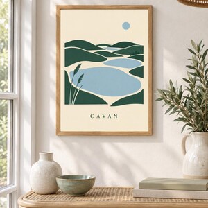 May include: A framed wall art print featuring a stylized landscape with dark green rolling hills, light blue lakes, and a small blue sun against a cream background. The word CAVAN is printed in black capital letters at the bottom of the artwork.