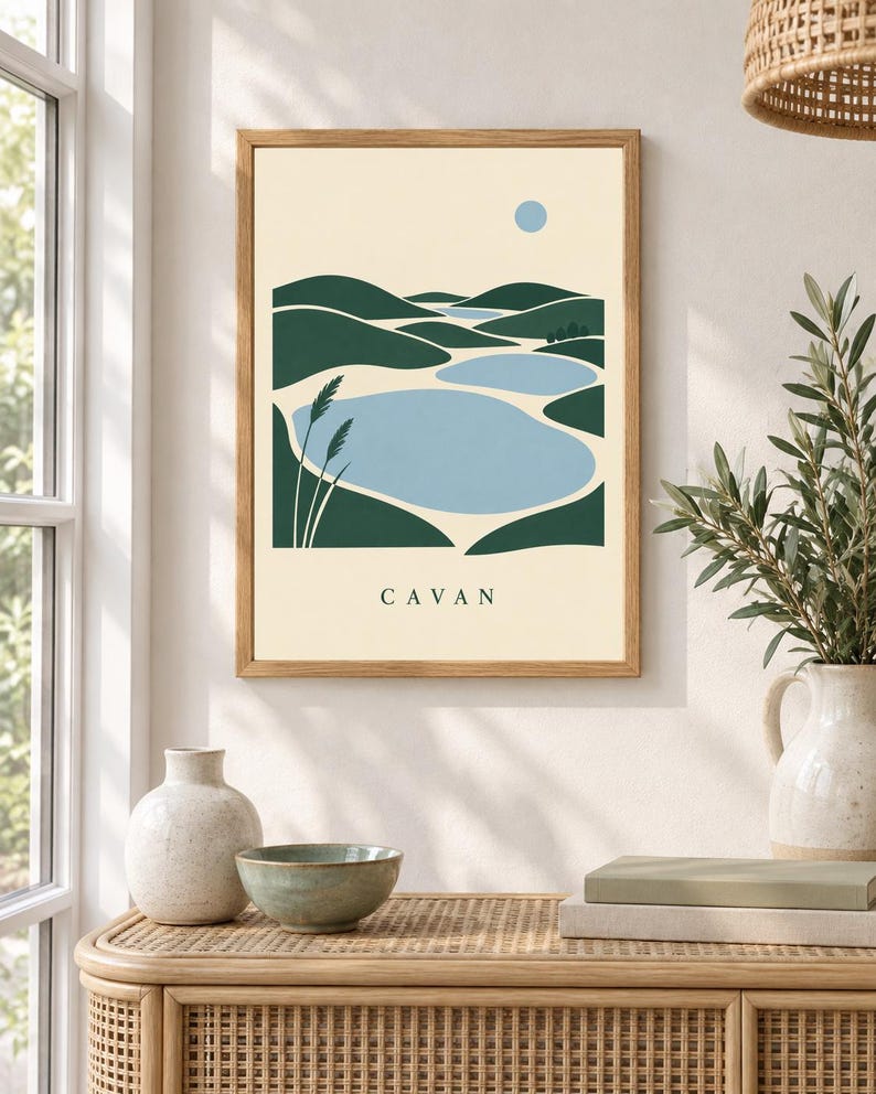 May include: A framed wall art print featuring a stylized landscape with dark green rolling hills, light blue lakes, and a small blue sun against a cream background. The word CAVAN is printed in black capital letters at the bottom of the artwork.