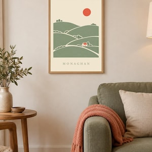 May include: A framed wall art print featuring a stylised landscape of rolling green hills, a small white house, and a red sun against a cream background. The word MONAGHAN is printed in black capital letters at the bottom of the illustration.