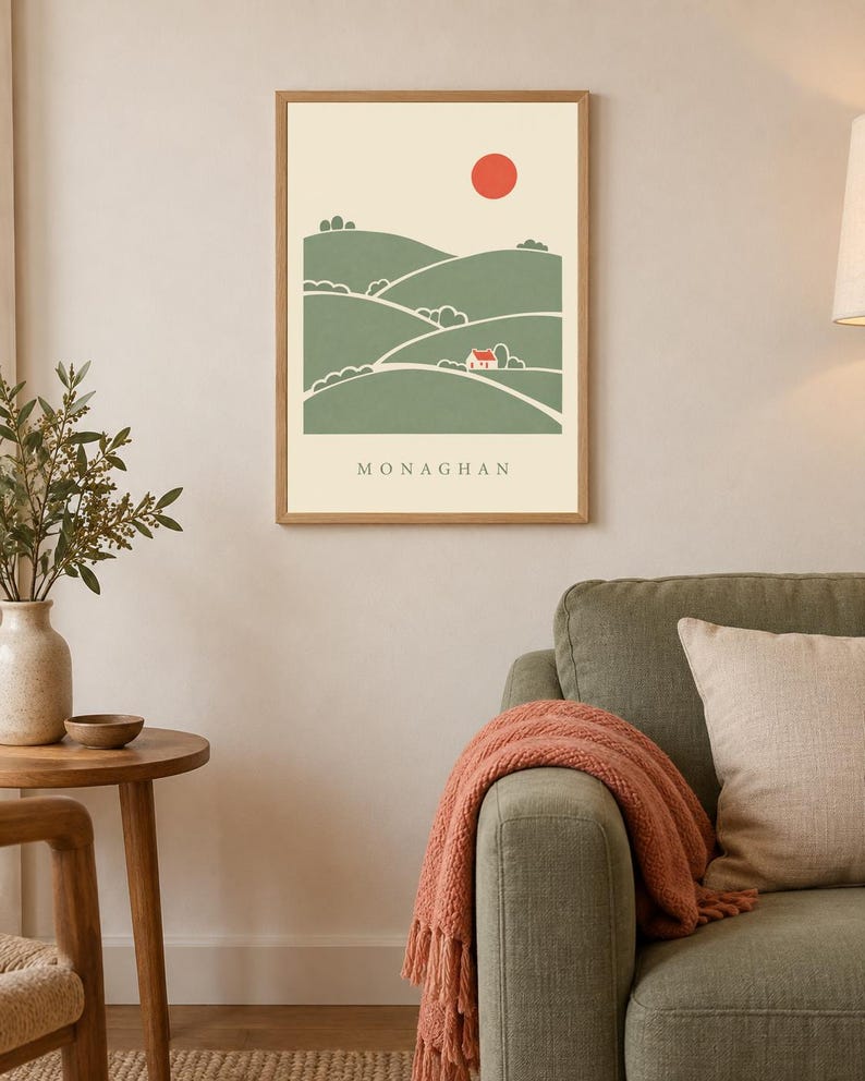 May include: A framed wall art print featuring a stylised landscape of rolling green hills, a small white house, and a red sun against a cream background. The word MONAGHAN is printed in black capital letters at the bottom of the illustration.