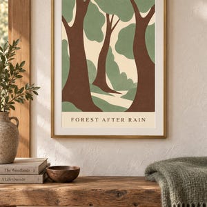 May include: A framed wall art print featuring a stylized forest scene with brown tree trunks and sage green foliage against a cream background. The text FOREST AFTER RAIN is printed in black at the bottom of the illustration within the frame.