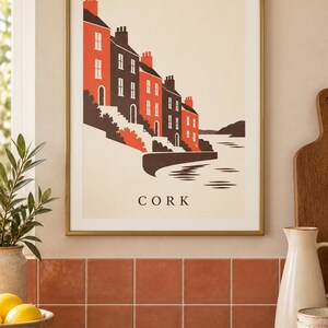 Op de afbeelding: Een ingelijste kunstdruk met een gestileerde illustratie van huizen langs het water in Cork. De afbeelding gebruikt een palet van rood, donkergrijs en cr&egrave;me, met het woord CORK in zwarte hoofdletters onderaan in het midden gedrukt.