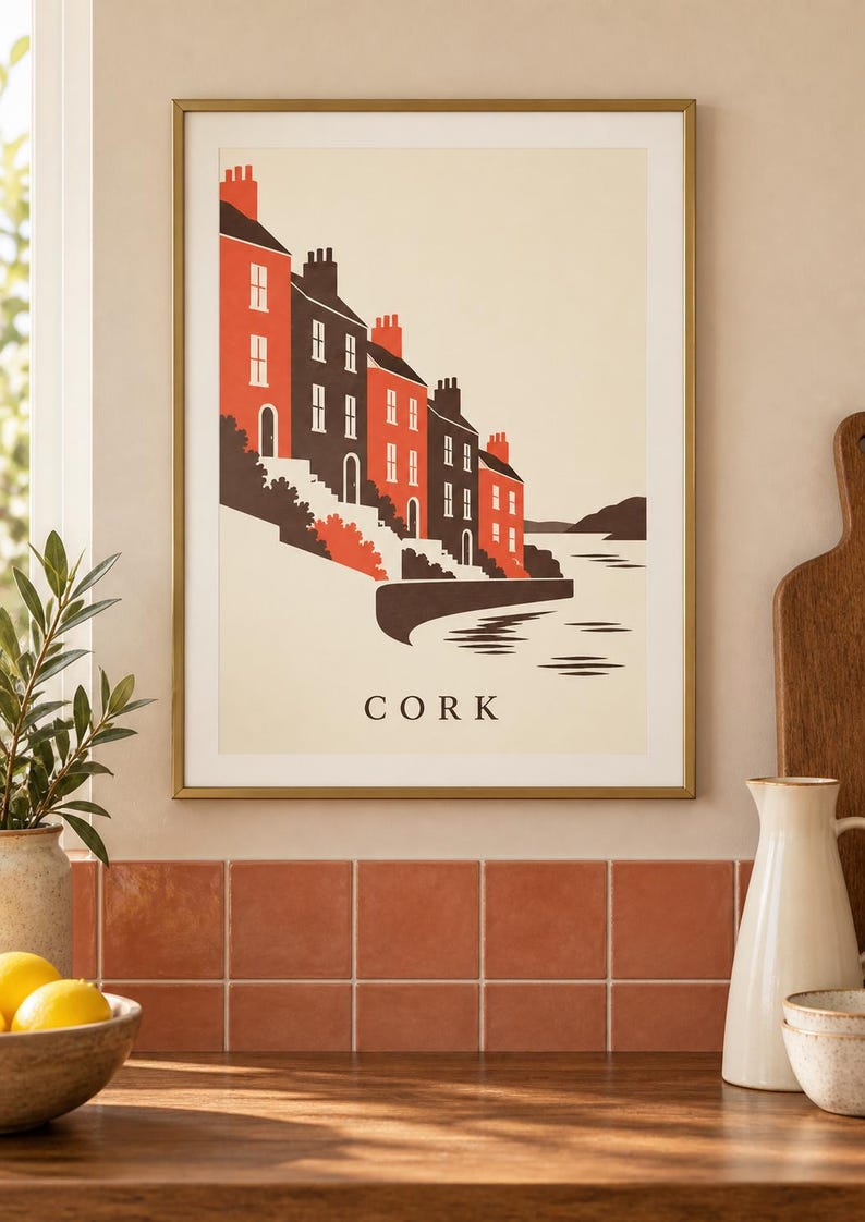 Op de afbeelding: Een ingelijste kunstdruk met een gestileerde illustratie van huizen langs het water in Cork. De afbeelding gebruikt een palet van rood, donkergrijs en cr&egrave;me, met het woord CORK in zwarte hoofdletters onderaan in het midden gedrukt.