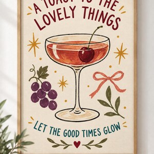 May include: A framed wall art print featuring a cocktail glass with a cherry, a cluster of purple grapes, a pink ribbon, and gold star accents. The text reads A TOAST TO THE LOVELY THINGS and LET THE GOOD TIMES GLOW, with a small red heart at the bottom.