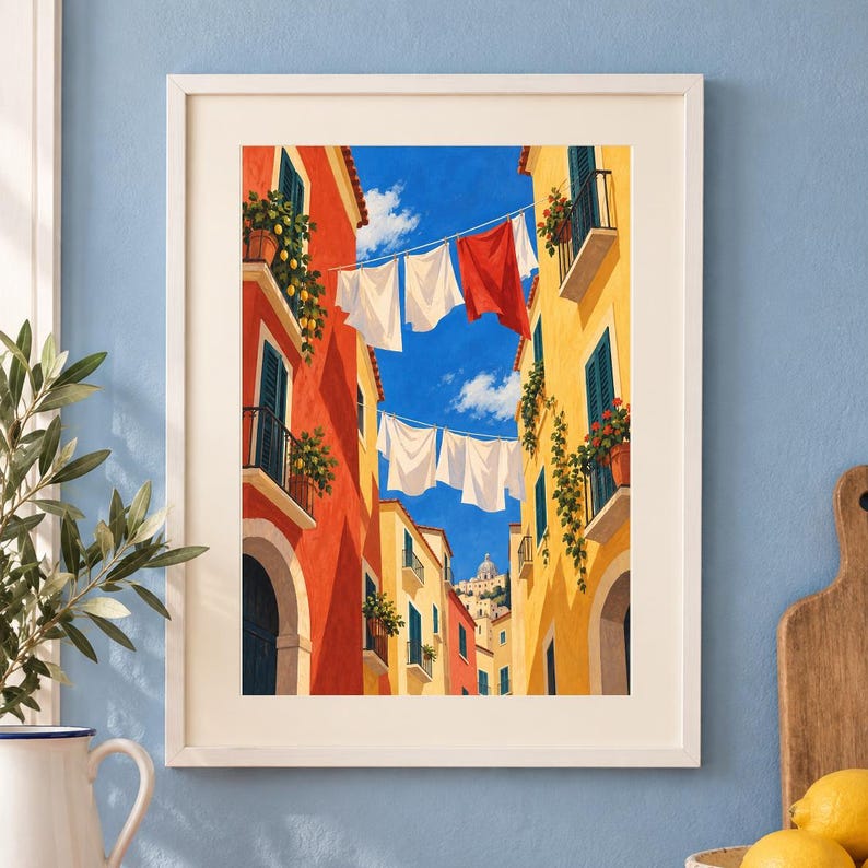Op de afbeelding: Een ingelijste kunstdruk toont een smalle straat tussen hoge rode en gele gebouwen onder een helderblauwe lucht. Wasgoed hangt aan lijnen tussen de gebouwen, waaronder witte en rode stoffen. Potplanten en citroenbomen sieren de balkons.