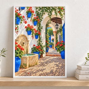 May include: A framed wall art print depicting a sunlit Mediterranean courtyard with a stone fountain, vibrant red geraniums in bright blue ceramic pots, climbing green vines, and a cobblestone path leading through a white arched stone entryway.