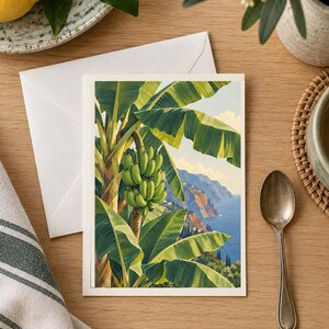 May include: A rectangular greeting card featuring a vibrant illustration of a banana tree with a large bunch of green bananas in the foreground, set against a scenic coastal landscape with blue ocean waters and cliffs under a clear sky.