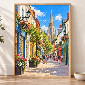 Op de afbeelding: Een ingelijste wandkunstprint met een zonnig Europees straatbeeld met kleurrijke winkelpuien, hangende bloemenmanden en voetgangers die over een geplaveid pad naar een hoge stenen kerktoren lopen onder een helderblauwe lucht.