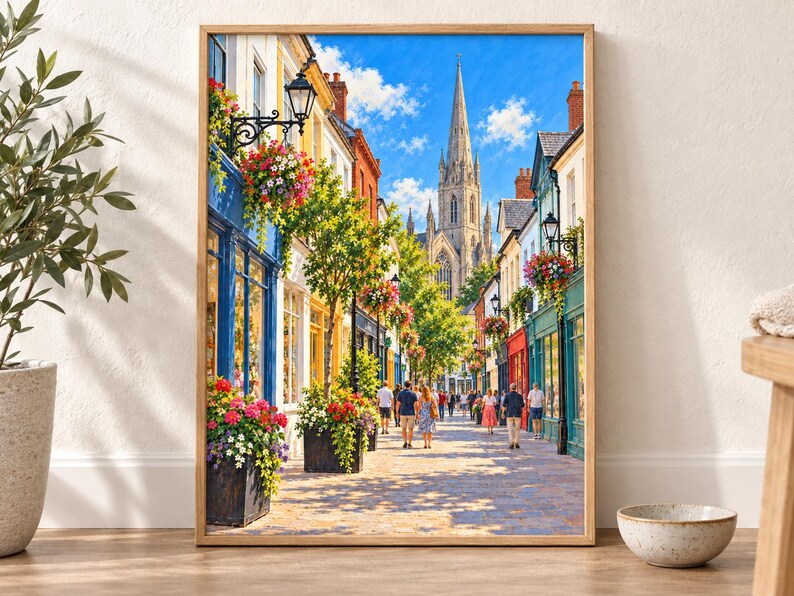 Op de afbeelding: Een ingelijste wandkunstprint met een zonnig Europees straatbeeld met kleurrijke winkelpuien, hangende bloemenmanden en voetgangers die over een geplaveid pad naar een hoge stenen kerktoren lopen onder een helderblauwe lucht.