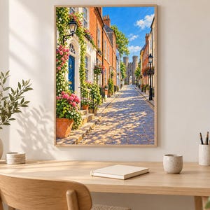 May include: A framed wall art print depicting a cobblestone street lined with brick buildings, vibrant pink and white flowers in terracotta pots, climbing greenery, a blue door, and a stone castle gatehouse under a bright blue sky.
