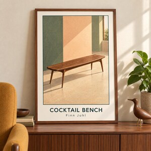 May include: A framed art print featuring a wooden bench with tapered legs in a room with green and peach walls. The text COCKTAIL BENCH and Finn Juhl is printed in black at the bottom of the off-white background within the frame.