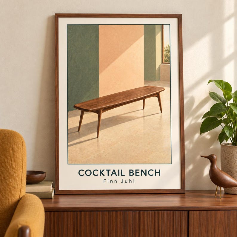 May include: A framed art print featuring a wooden bench with tapered legs in a room with green and peach walls. The text COCKTAIL BENCH and Finn Juhl is printed in black at the bottom of the off-white background within the frame.