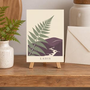 May include: A rectangular print on a small wooden easel featuring a green fern illustration over a dark purple landscape with a winding white river and the text LAOIS printed in black at the bottom against a cream-coloured background.