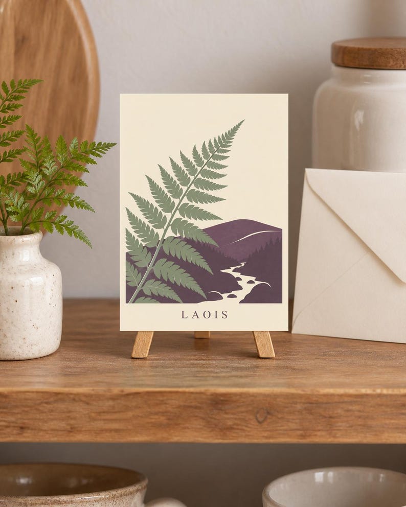 May include: A rectangular print on a small wooden easel featuring a green fern illustration over a dark purple landscape with a winding white river and the text LAOIS printed in black at the bottom against a cream-coloured background.
