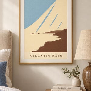 May include: A framed wall art print titled ATLANTIC RAIN featuring a minimalist landscape illustration with a light blue sky, cream-colored clouds, and dark brown coastal cliffs. The artwork is enclosed in a thin gold-toned metal frame.