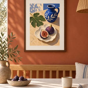 May include: A framed art print featuring a still life illustration of a blue ceramic pitcher with white leaf patterns, a single green fig leaf, and two purple figs, one whole and one halved, resting on a white scalloped plate on a tan surface.