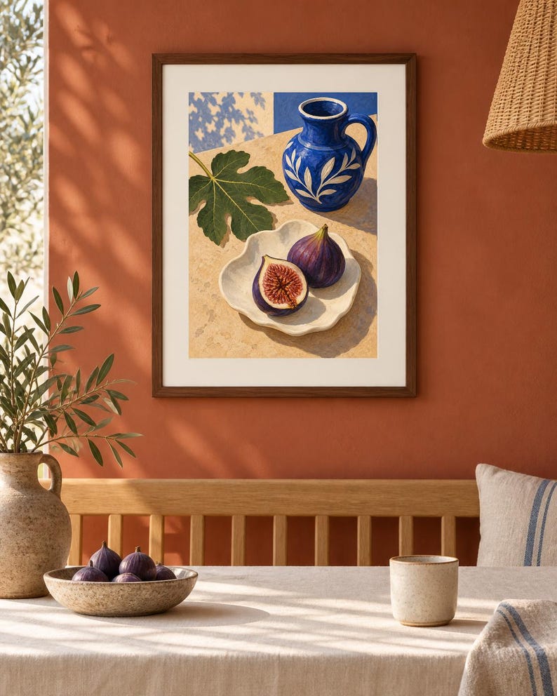 May include: A framed art print featuring a still life illustration of a blue ceramic pitcher with white leaf patterns, a single green fig leaf, and two purple figs, one whole and one halved, resting on a white scalloped plate on a tan surface.