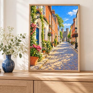May include: A framed wall art print depicting a cobblestone street lined with colorful brick buildings, climbing green vines, and pink flowers. A blue door is visible on the left, and a stone archway stands in the distance under a bright blue sky.